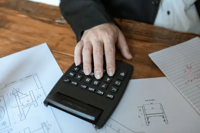 Teknik çizim üzerinde hesap makinesiyle maliyet hesabı yapan bir kişi